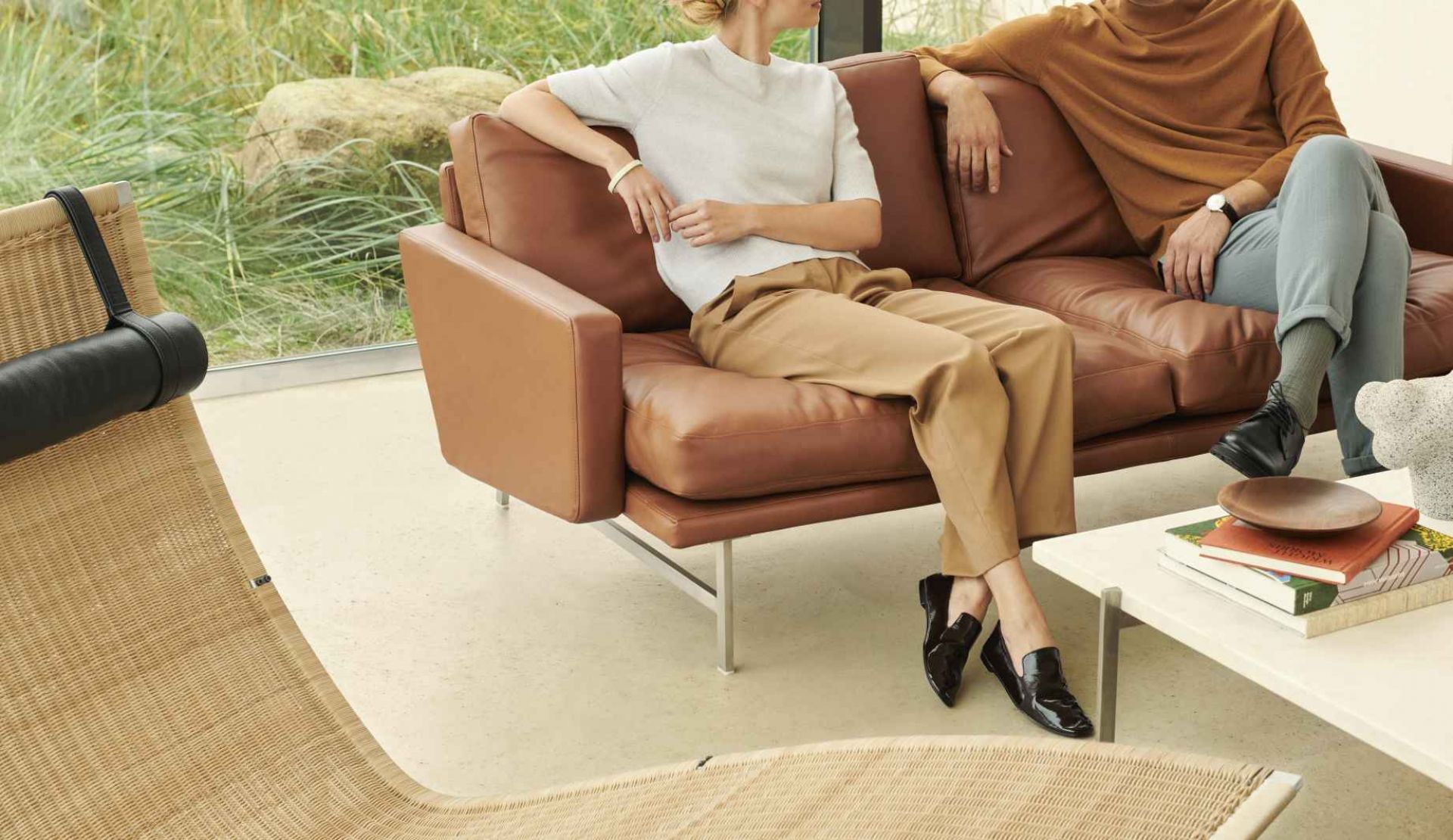 Zwei Personen entspannen auf einem braunen Fritz Hansen PK24 Ledersofa mit Stahlgestell in einem hellen Wohnraum.