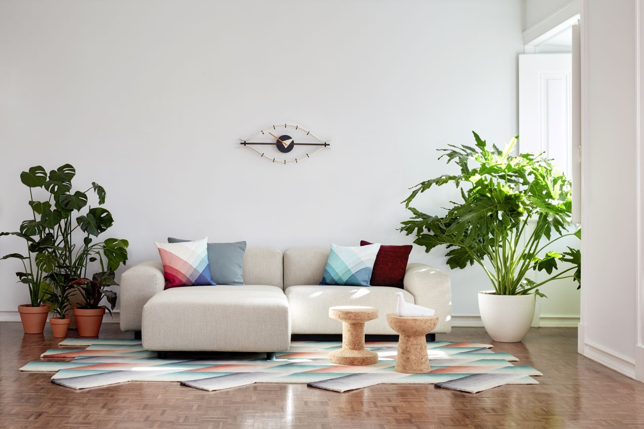 Wohnzimmer mit Vitra Eye Clock Wanduhr über Sofa, Zimmerpflanzen und geometrischem Teppich für modernes Wohnen.