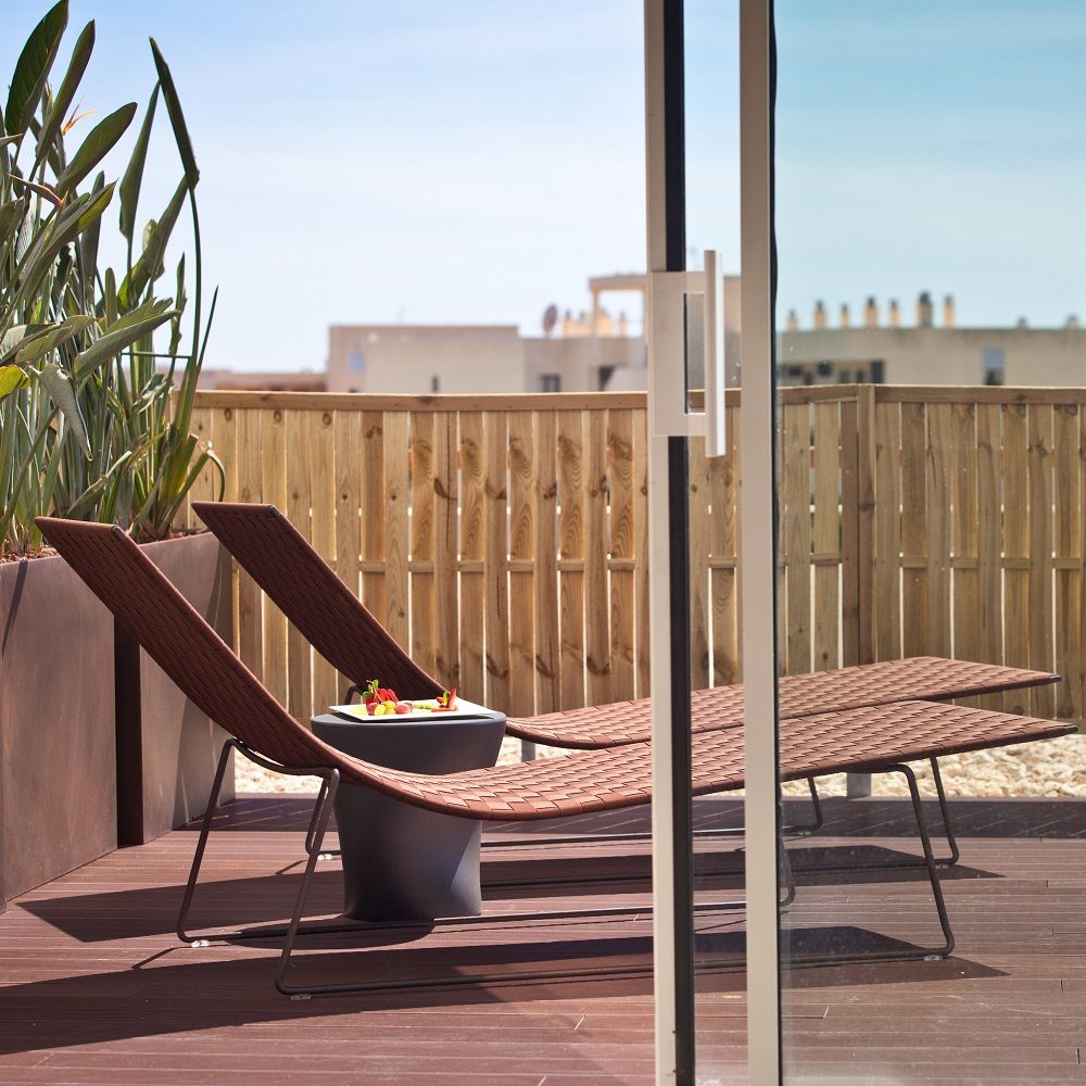 Andreu World Trenza Outdoor Liege auf Holzterrasse mit Getränkehalter und Blick auf Holzzaun.