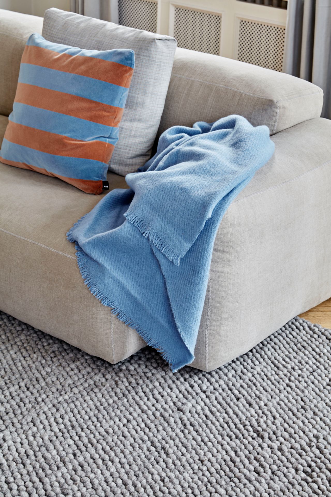 Beige Mags Soft Sofa mit blauer Decke und gestreiftem Kissen auf grauem Teppich.