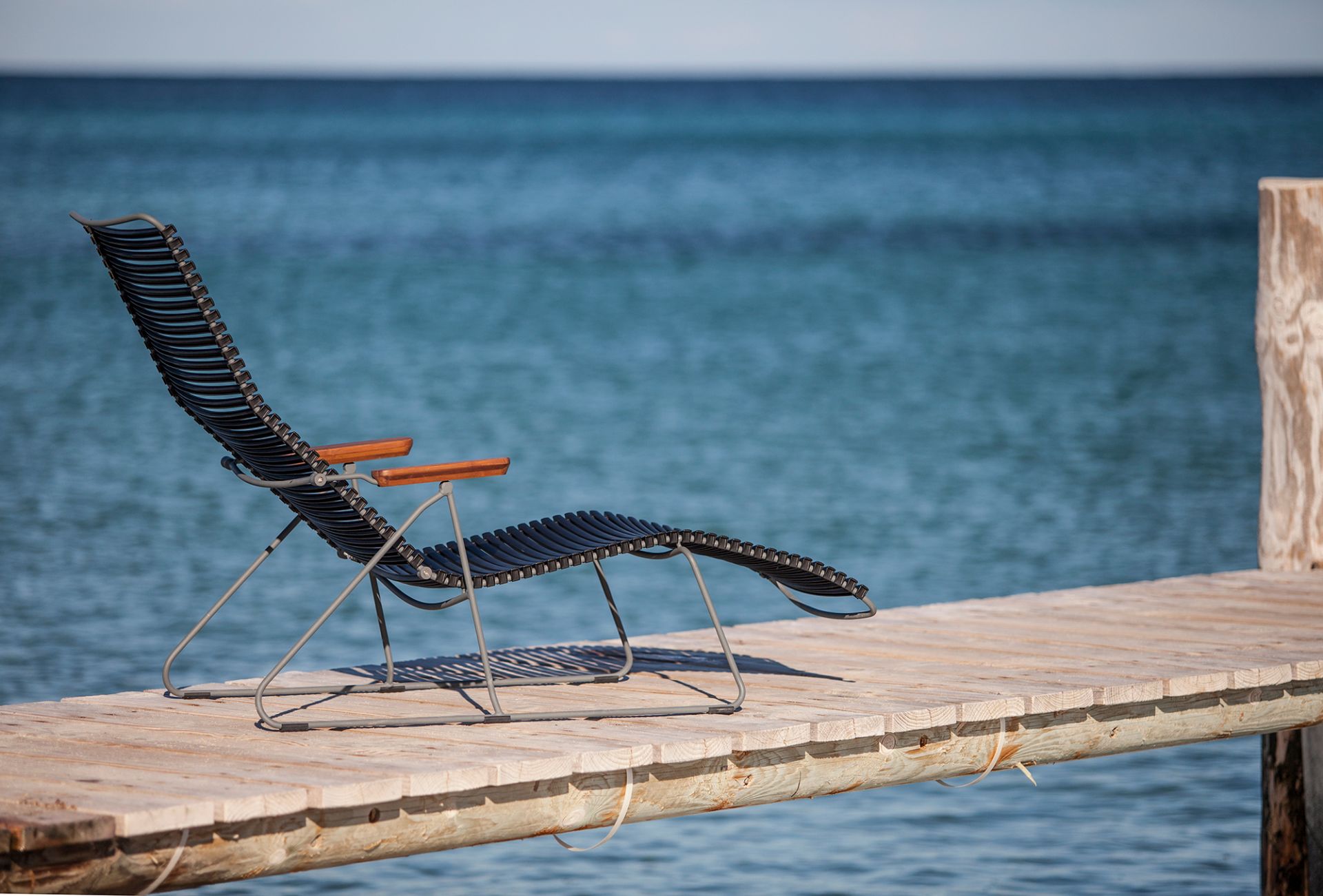 Click Sunlounger Outdoor Sonnenliege von Houe in Dunkelgrau auf einem Holzsteg mit Meerblick.