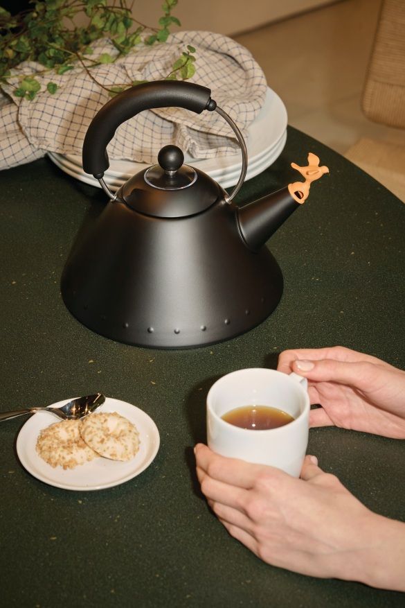 Schwarzer Alessi 9093 BB Wasserkessel mit Vogelpfeife, Tasse Tee und Gebäck auf einem Tisch.