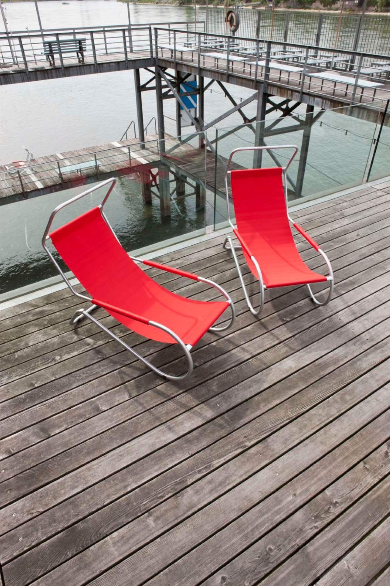 Zwei rote Lido Liegen mit silbernem Gestell auf einer Holzterrasse am Wasser.