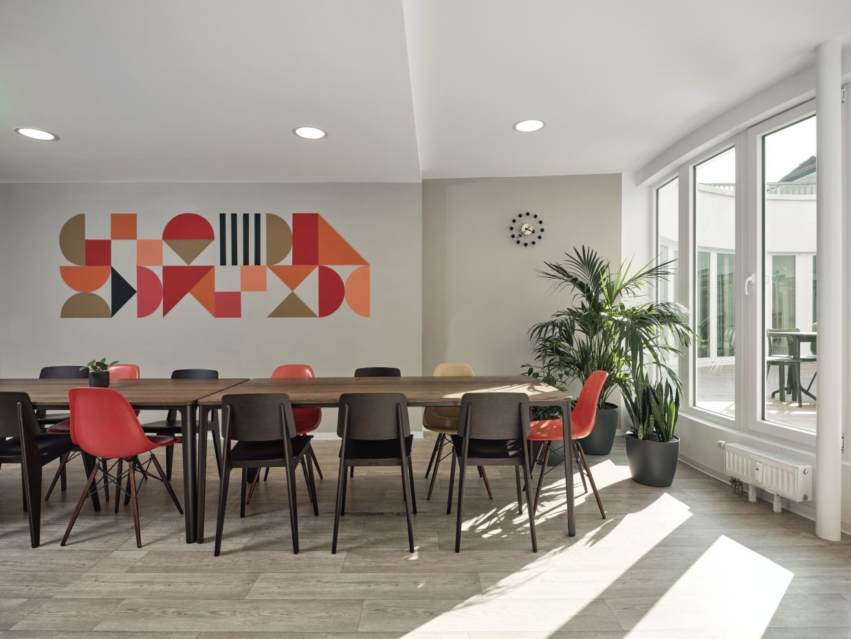 Modernes Besprechungszimmer mit großem Holztisch, farbigen Stühlen und geometrischer Wandkunst für inspirierende Meetings.