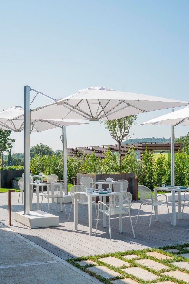 Außenbereich mit weißen Emu Nova Tischen und Stühlen unter großen Sonnenschirmen auf einer Terrasse.