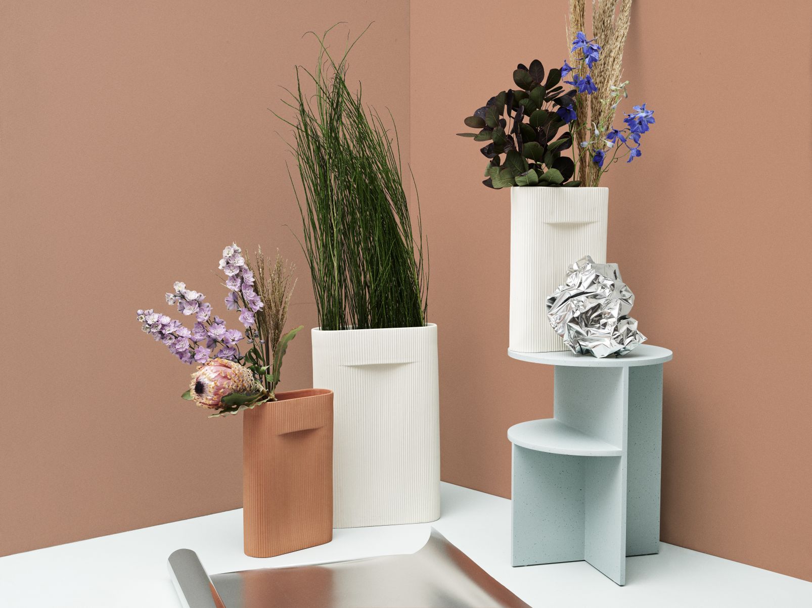 Drei dekorative Vasen, darunter die weiße Muuto Ridge Vase, gefüllt mit Blumen und Gräsern, auf Podest.