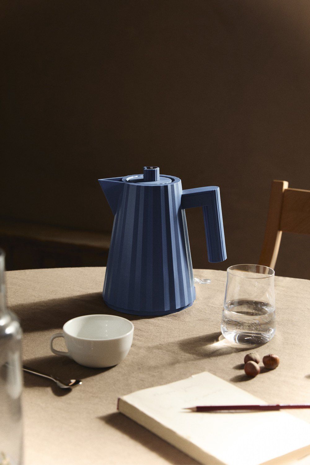 Blauer Alessi Plissé Wasserkocher auf Tisch mit Tasse, Glas und Notizbuch.