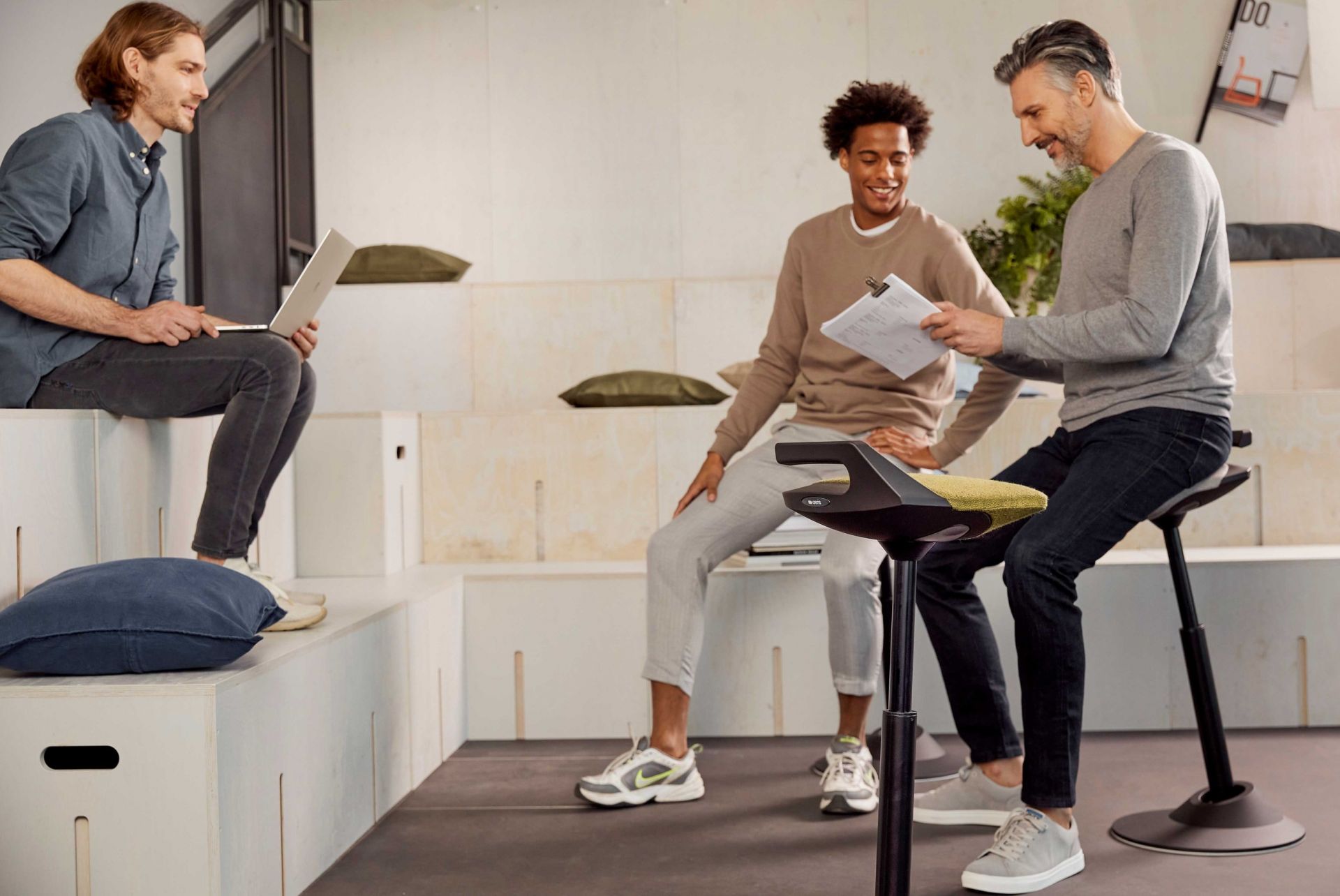 Drei Personen in einem Büro, eine Person sitzt auf einem Steh-Sitz-Hocker mit gelbem Sitz.