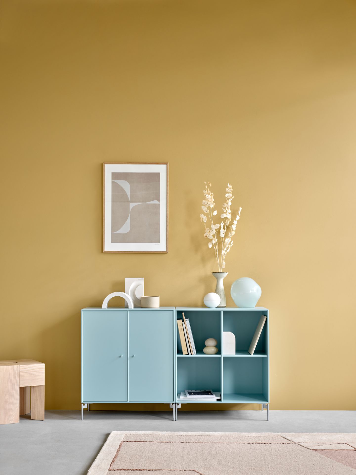 Helles, blaues Montana Sideboard mit Fächern und Deko-Elementen vor einer ockerfarbenen Wand.
