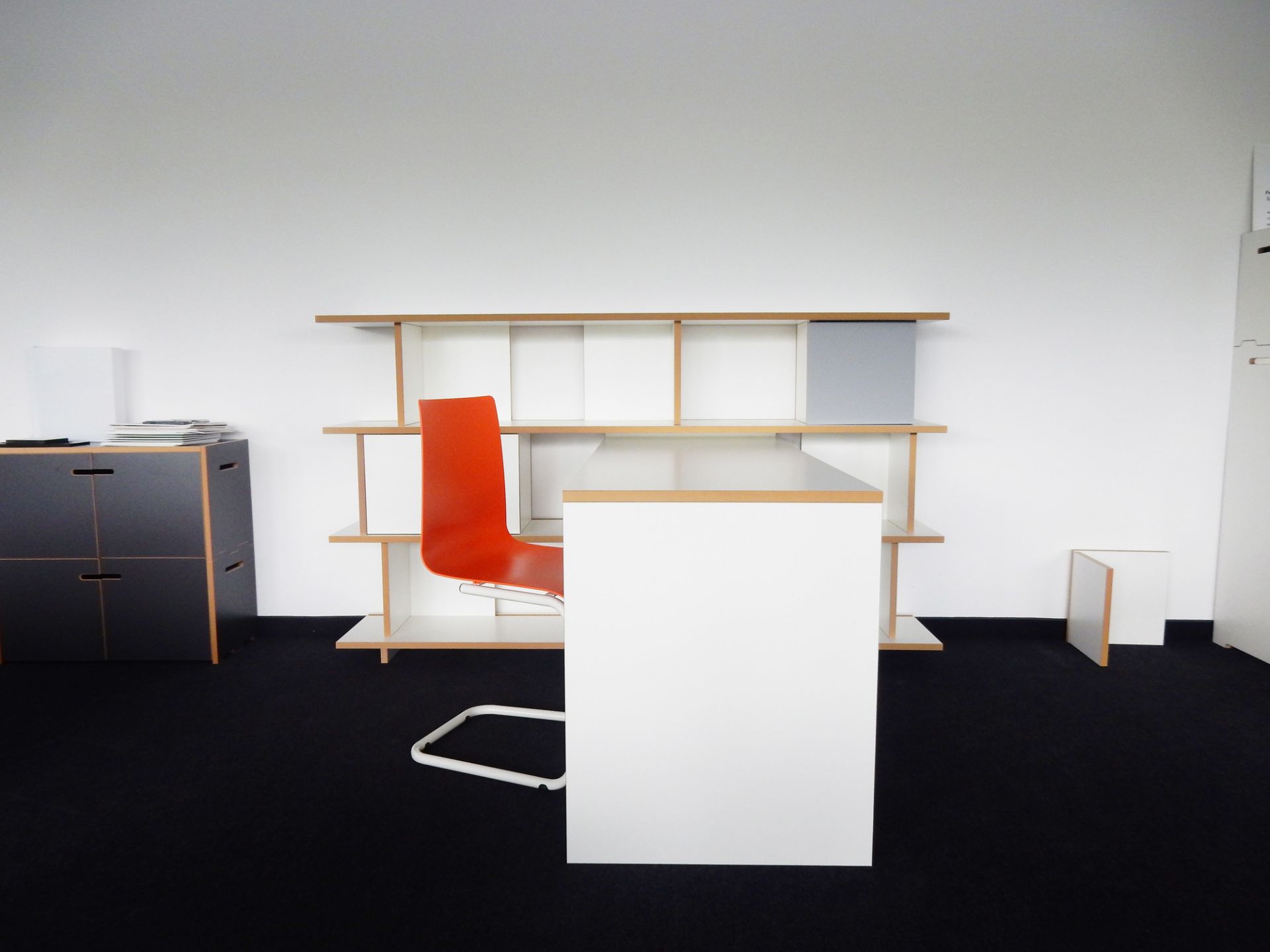 Tojo Anstelltisch für Stell Regal im modernen Büro mit orangefarbenem Stuhl und schwarzem Teppich.