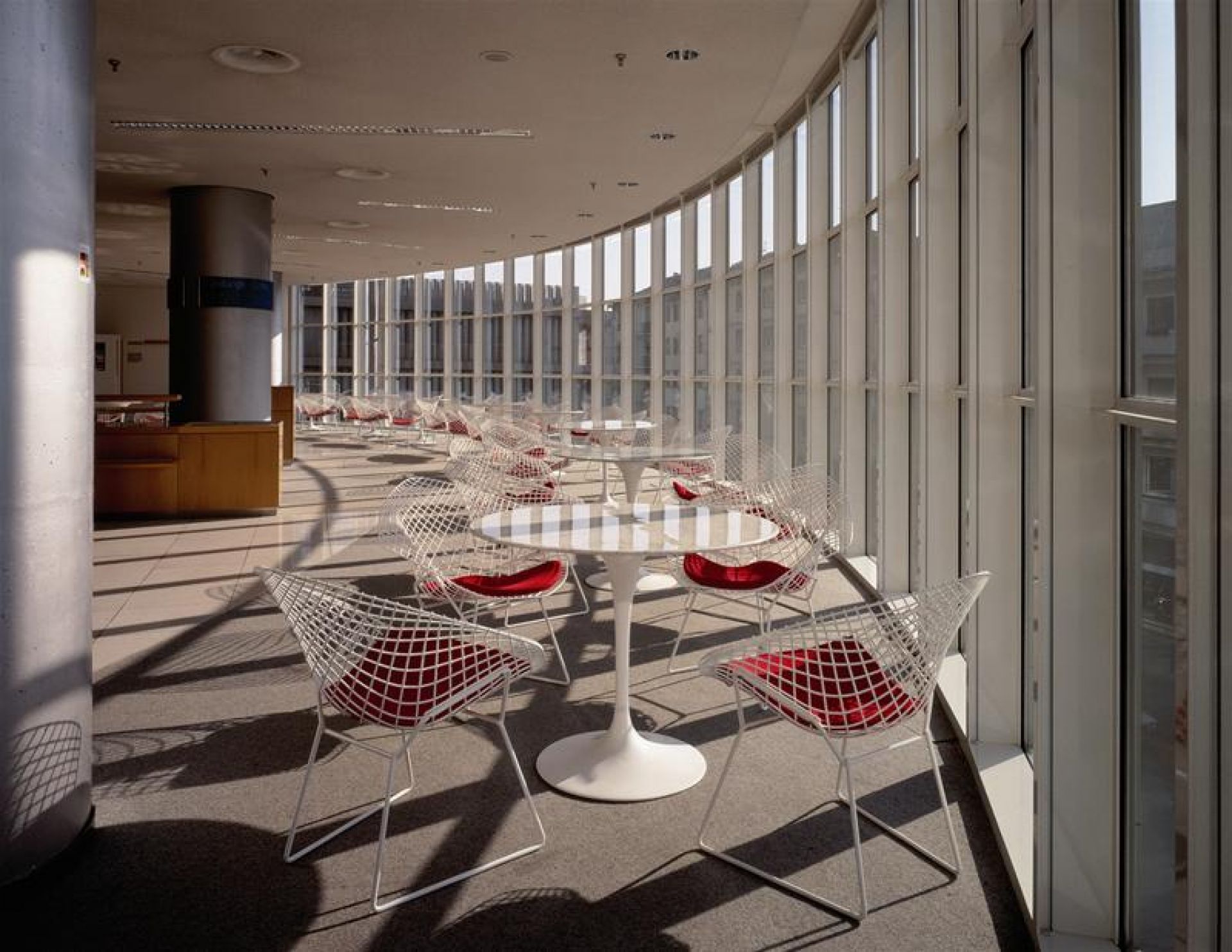 Helle Cafeteria mit weißen Saarinen Esstischen von Knoll International und Drahtstühlen mit roten Sitzkissen vor einer Fensterfront.