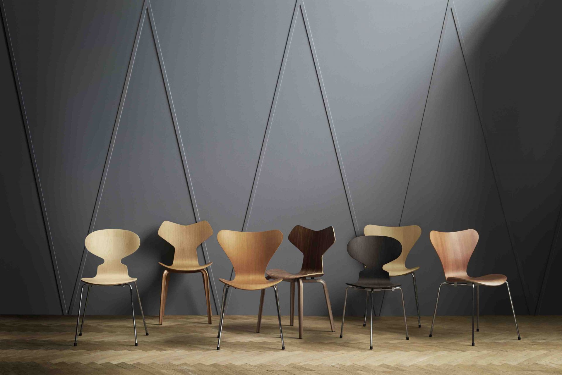 Eine Reihe von Fritz Hansen Grand Prix Stühlen aus Holz in verschiedenen Ausführungen vor grauer Wand.