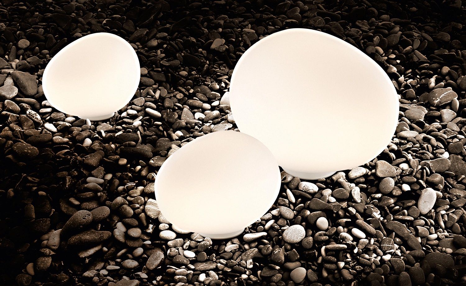 Foscarini Gregg Outdoor Leuchten: Drei eiförmige Bodenleuchten in Weiß auf dunklem Kiesstrand, stimmungsvolle Gartenbeleuchtung.