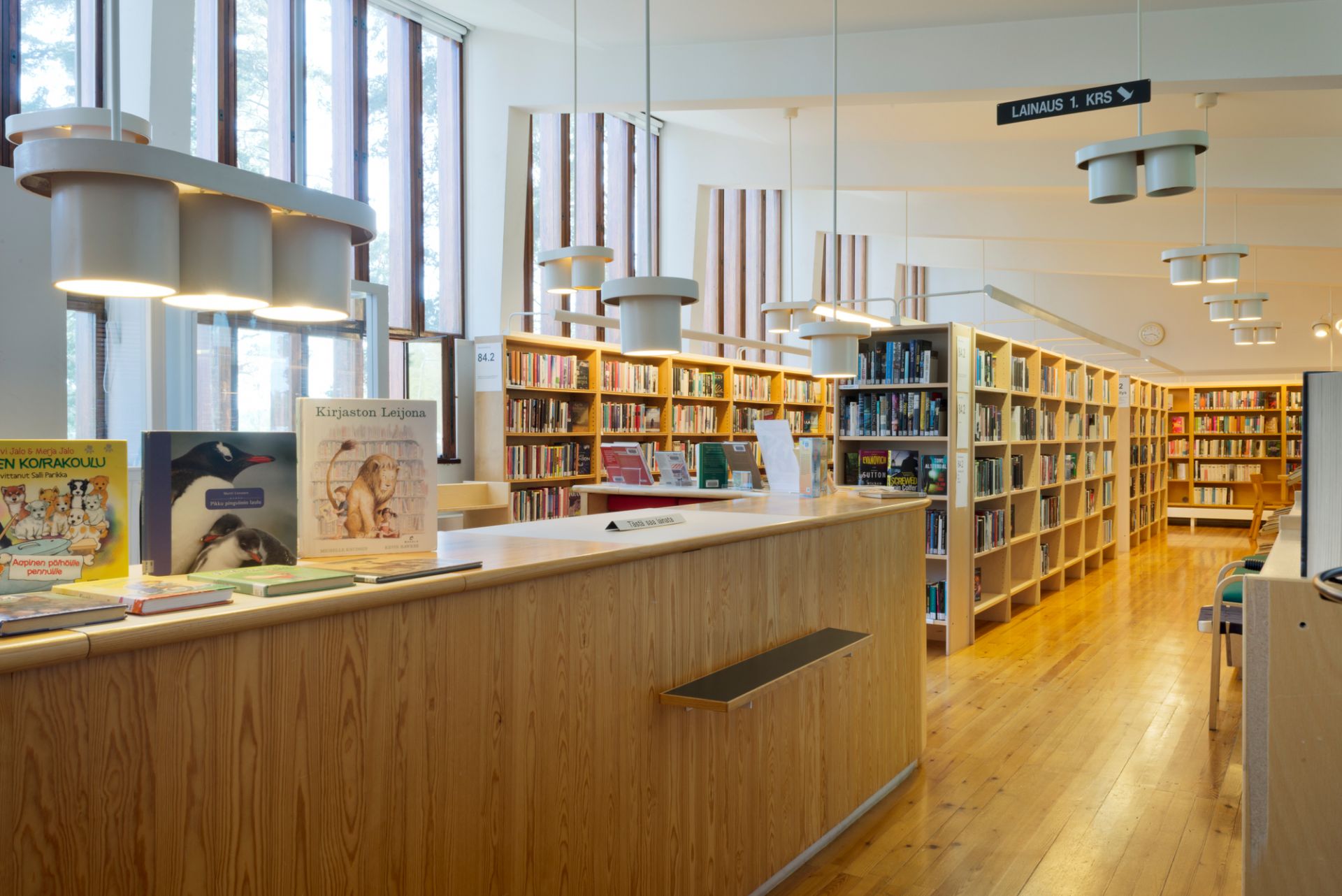 Helle Bibliothek mit Artek A201 Pendelleuchten, die über der hölzernen Ausleihtheke hängen.