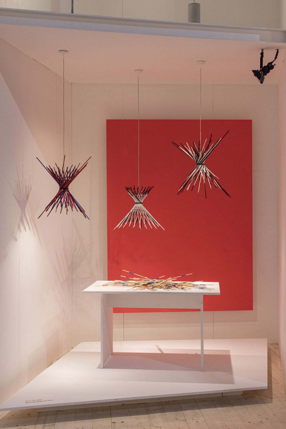 Drei Pic-A-Stic Pendelleuchten von Ingo Maurer in einem Ausstellungsraum mit rotem Hintergrund und weißem Tisch.
