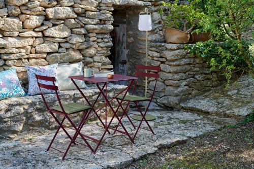Burgunderroter Fermob Bistro Tisch mit Stühlen im Freien vor einer Steinmauer. Outdoor Möbel für Garten und Balkon.