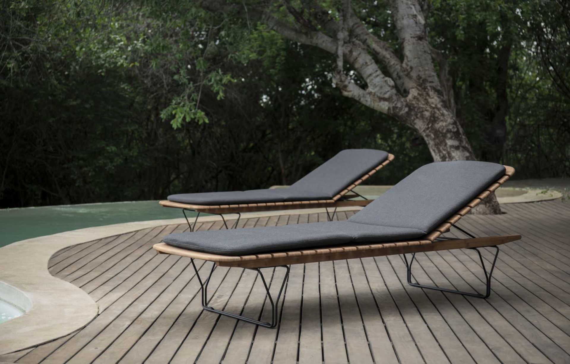 Zwei Houe Sonnenliegen mit grauen Molo Outdoor Kissen auf einer Holzterrasse neben einem Pool.