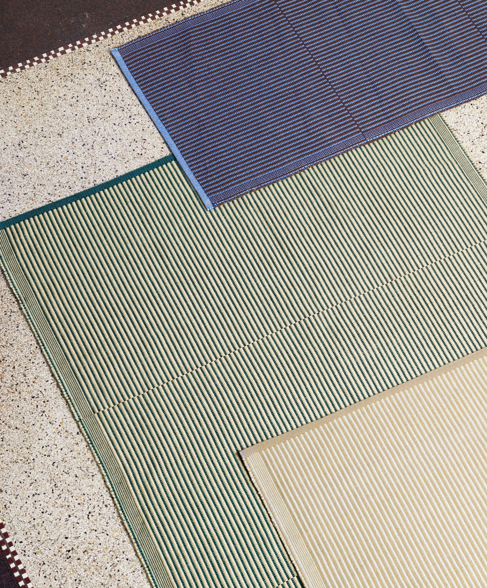 Nahaufnahme von drei gestreiften Hay Teppichen in Blau, Grün und Beige auf einem Terrazzoboden.