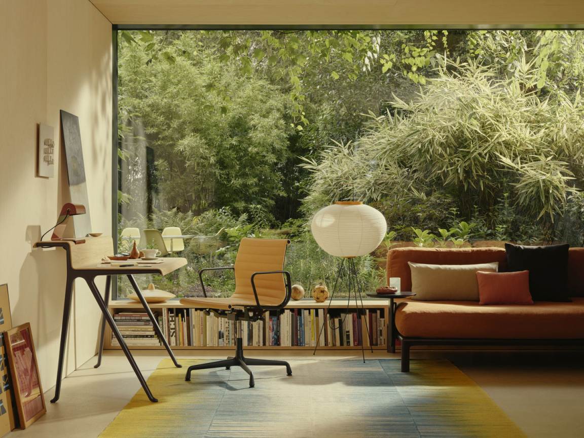 Heller Raum mit Vitra Courier Schreibtisch, Bürostuhl, Bücherregal und Sofa vor großem Fenster mit Gartenblick.