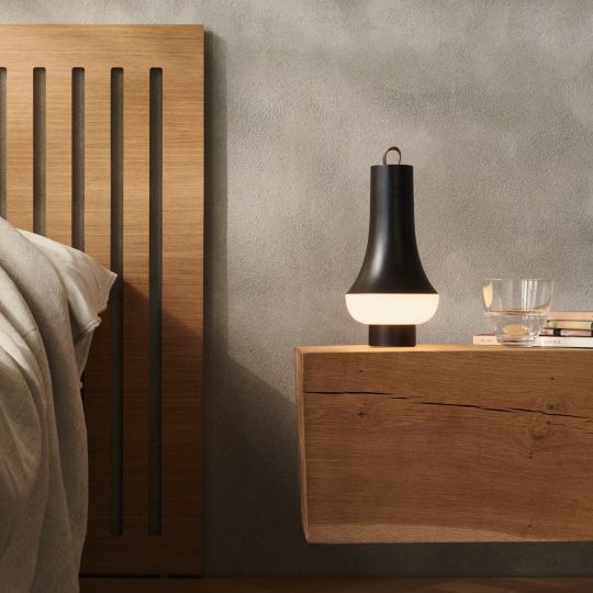 Tomoshi Portable Akkuleuchte von Louis Poulsen in Dunkelbraun auf einem Nachttisch neben einem Bett.