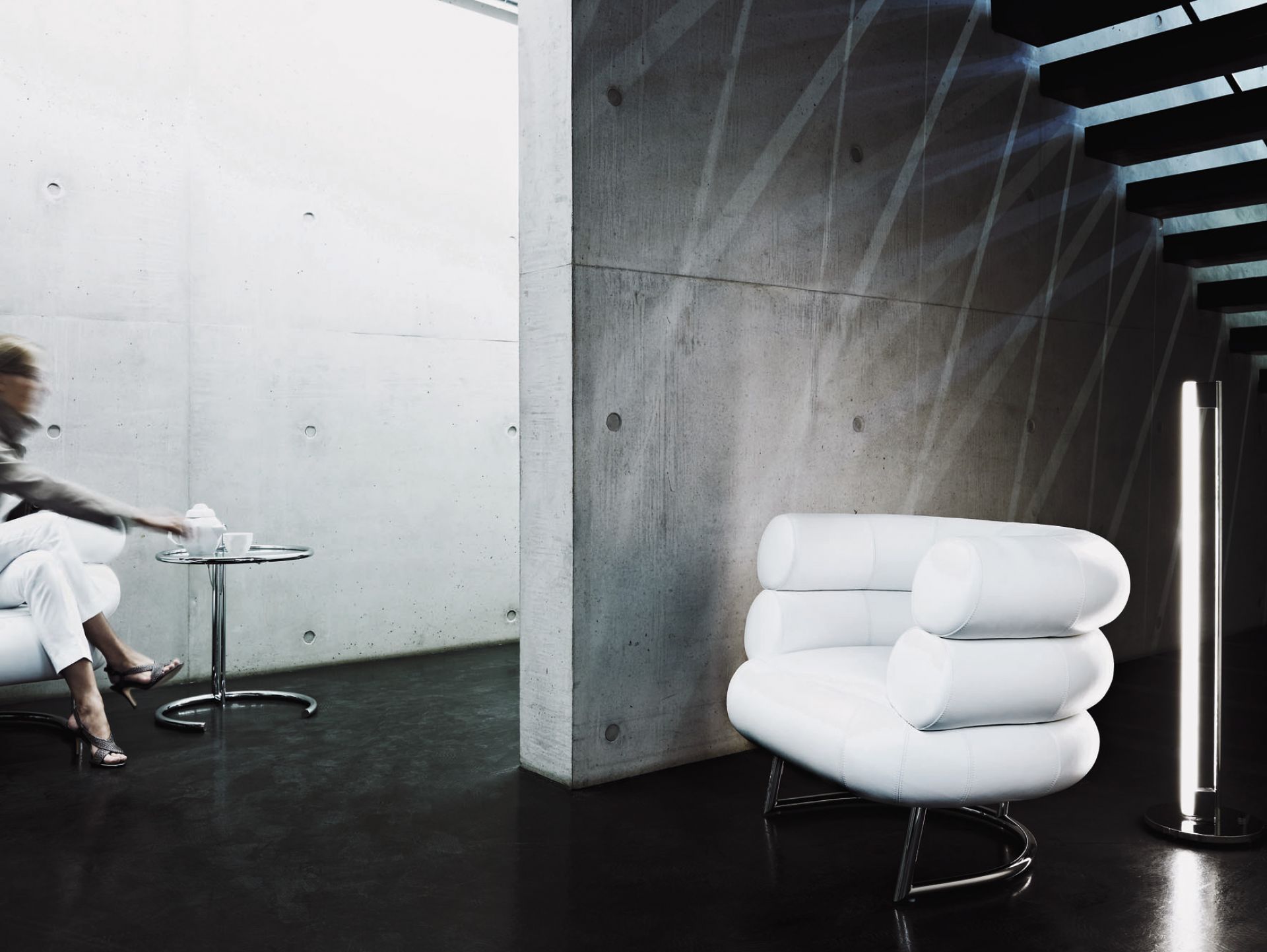 Moderner Wohnraum mit Adjustable Table E1027 Beistelltisch, Designklassiker mit Glasplatte und Stahlrohrgestell neben weißem Sessel.