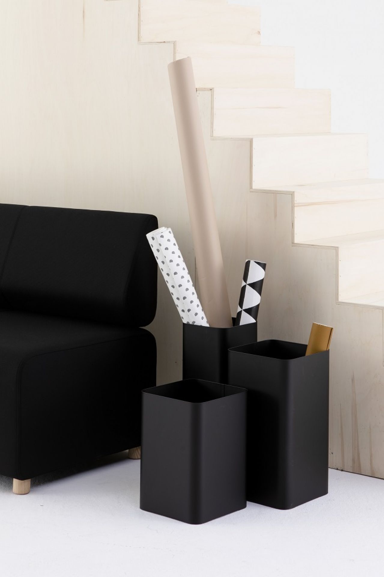 Drei schwarze Cascando Flow Papierkörbe unterschiedlicher Größe neben einem schwarzen Sofa und einer Holztreppe.