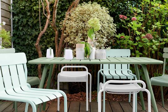 Hellgrüner Luxembourg Hocker von Fermob und passende Gartenmöbel auf einer Holzterrasse vor üppigem Grün.