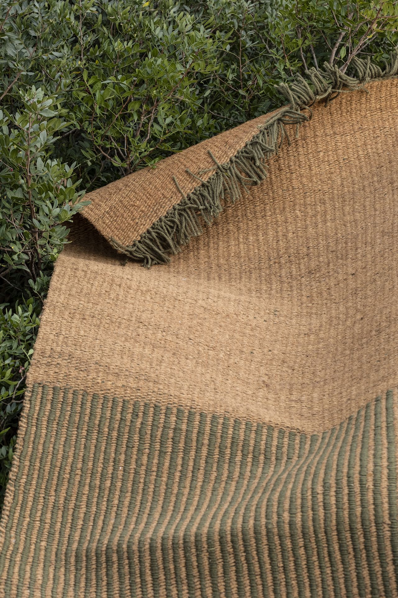 Nanimarquina Gartenteppich aus Naturfaser mit grünen Streifen und Fransen vor grünen Büschen.