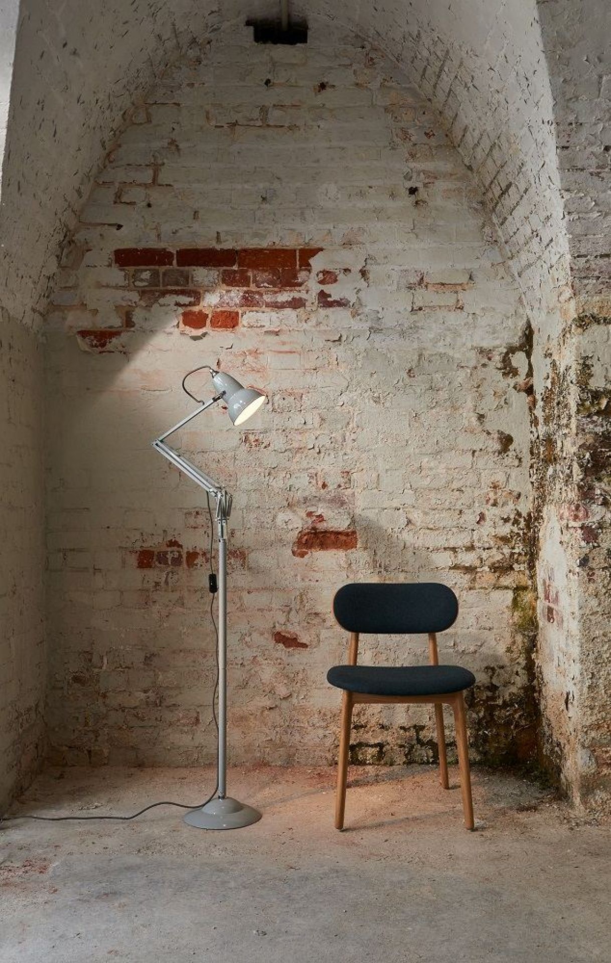 Anglepoise Original 1227 Stehleuchte in Taubengrau neben einem Stuhl vor Backsteinwand.