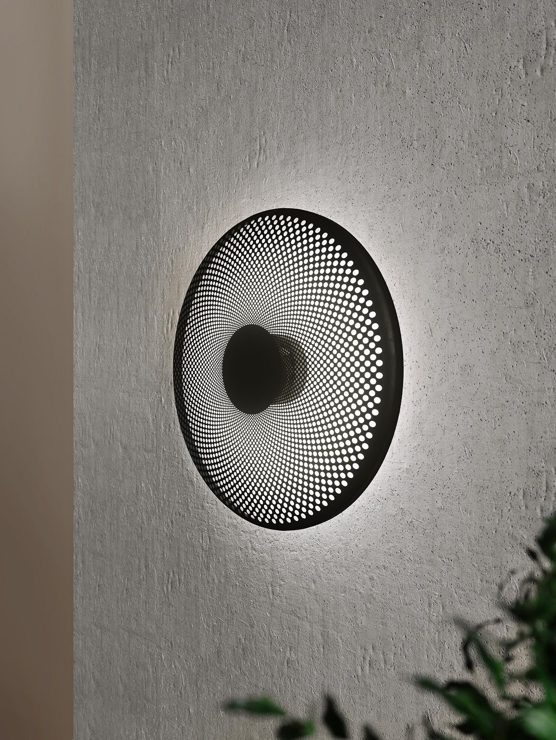 Glint Wandleuchte von Northern, runde, schwarze Designlampe mit Lochmuster an einer grauen Wand.