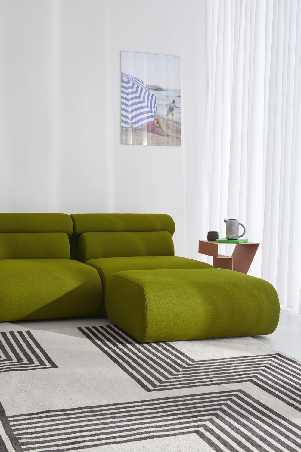 Grünes OLA Sofa mit passendem Pouf auf gemustertem Teppich, neben kleinem Beistelltisch und Vorhang.