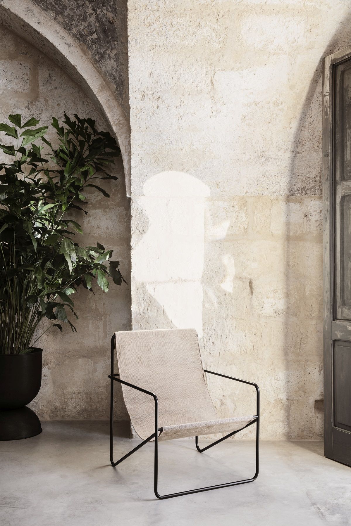 Desert Chair Loungesessel von Ferm Living mit beigem Stoffbezug und schwarzem Metallgestell vor Steinwand.