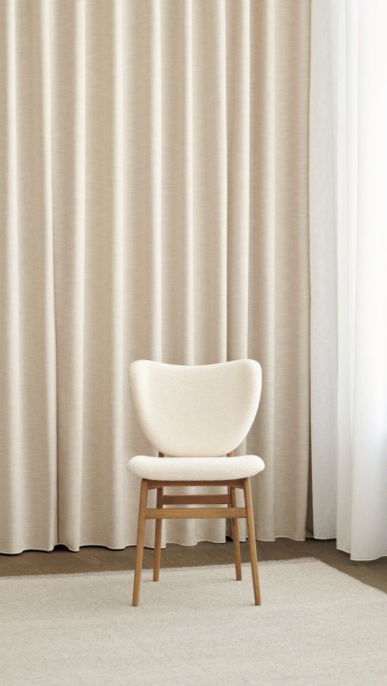 NORR11 Elephant Dining Chair, frontgepolstert, mit hellem Bouclé-Bezug und Holzbeinen vor beigen Vorhängen.