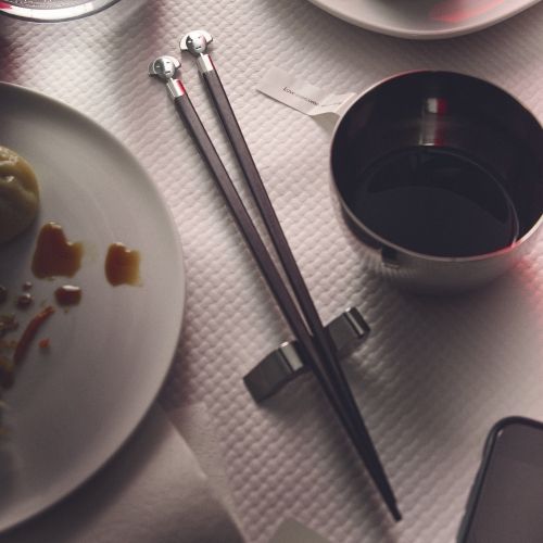 Alessi Anna Stick Essstäbchen Set auf gedecktem Tisch mit Sojasauce und Speisen.