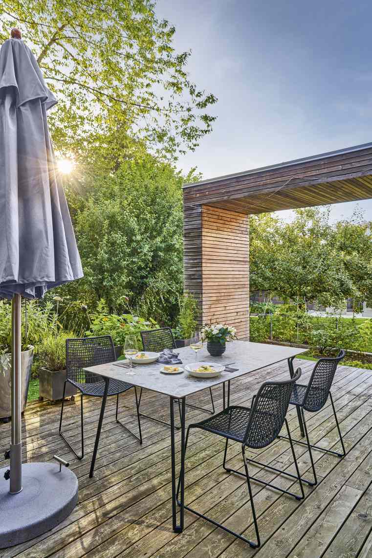 Weishäupl Slope Tisch auf Holzterrasse mit Stühlen, Sonnenschirm und Speisen für ein entspanntes Outdoor-Dinner.