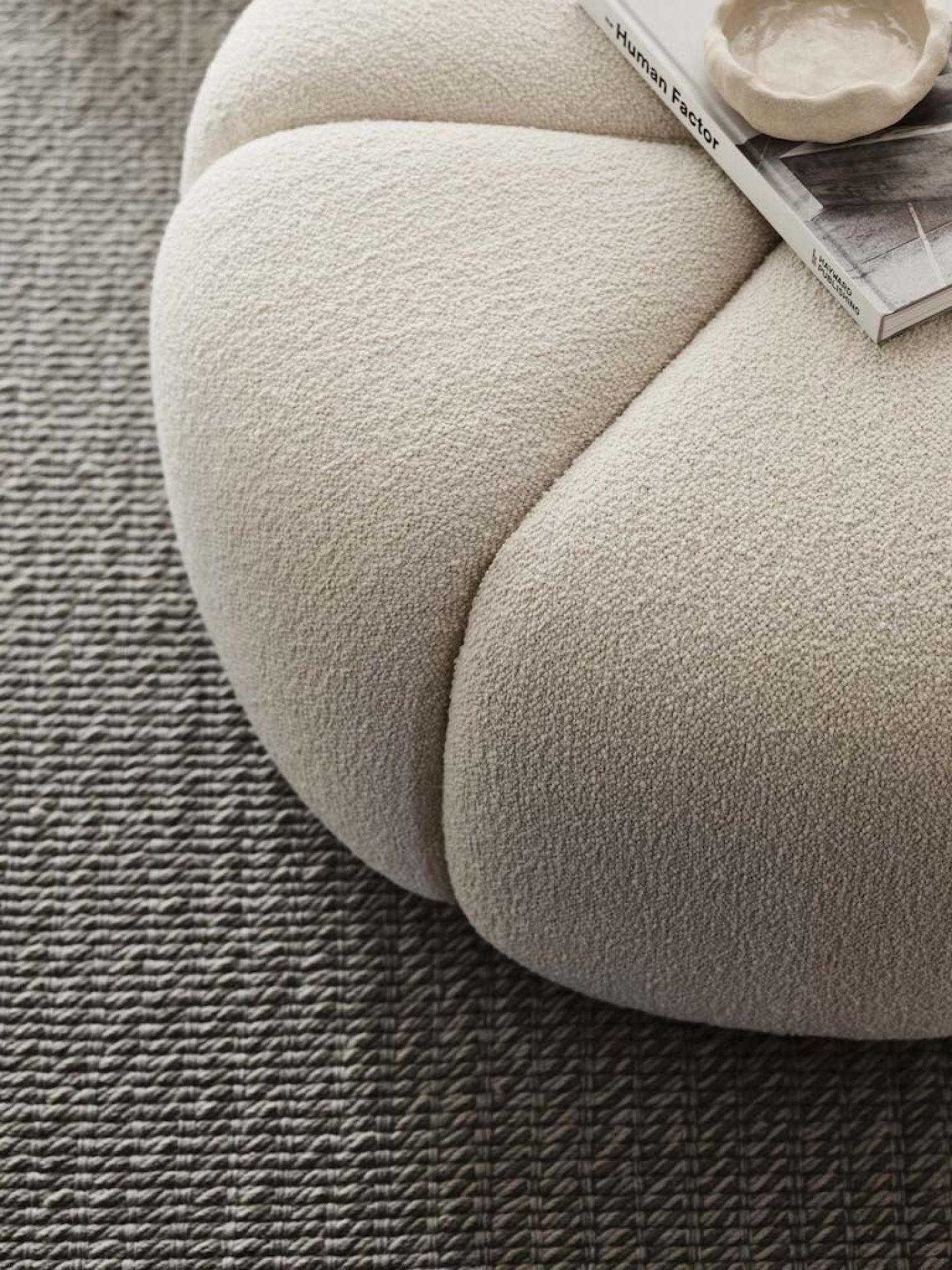Detailaufnahme: Off White Pouf Hocker mit Buch und Schale auf einem strukturierten Teppich.