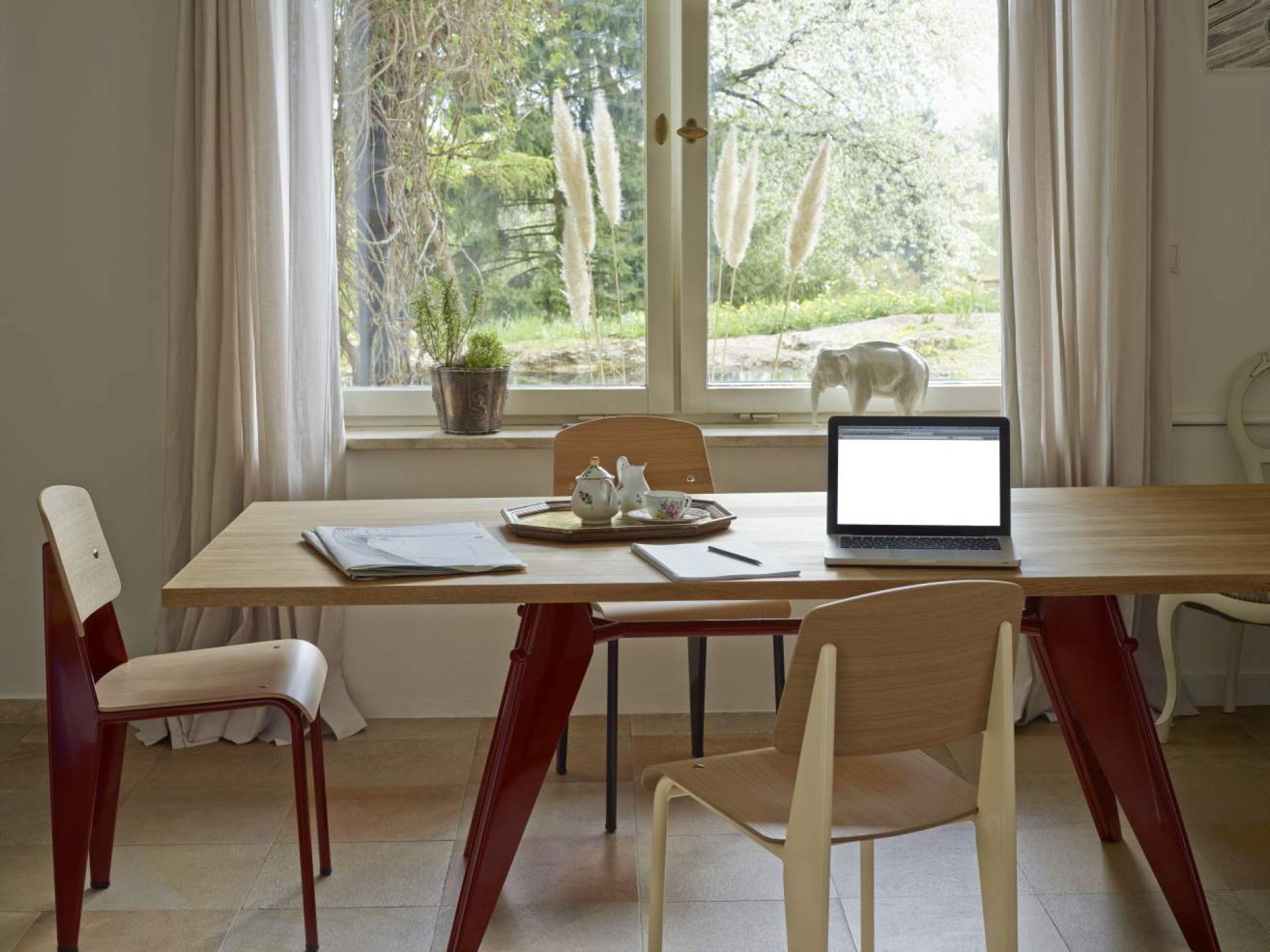 Vitra EM Table Holztisch mit Laptop, Geschirr und Stühlen vor Fenster mit Gartenblick.
