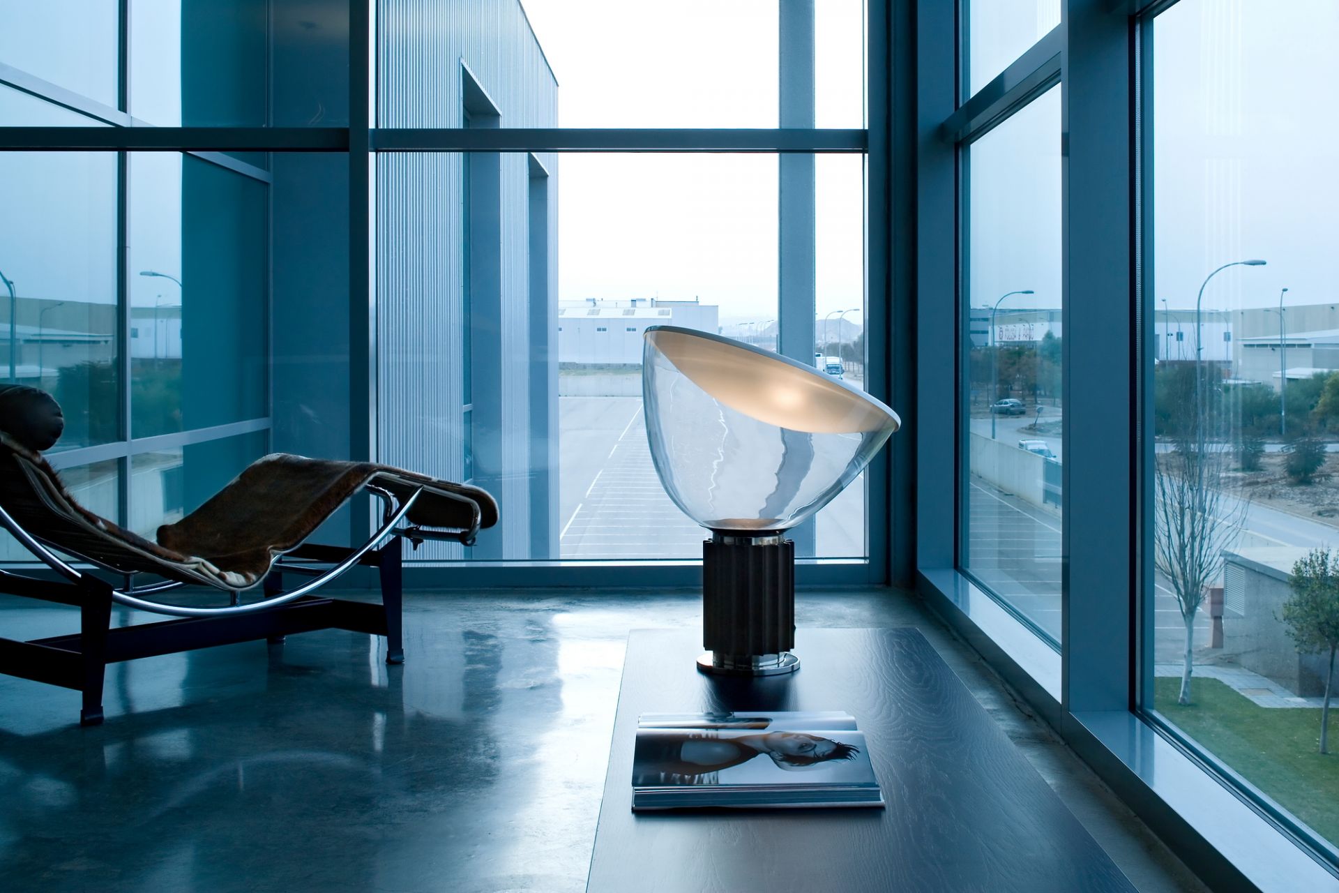 Taccia Tischleuchte von Flos im modernen Interieur mit Sessel und großem Fenster, stimmungsvolle Beleuchtung.