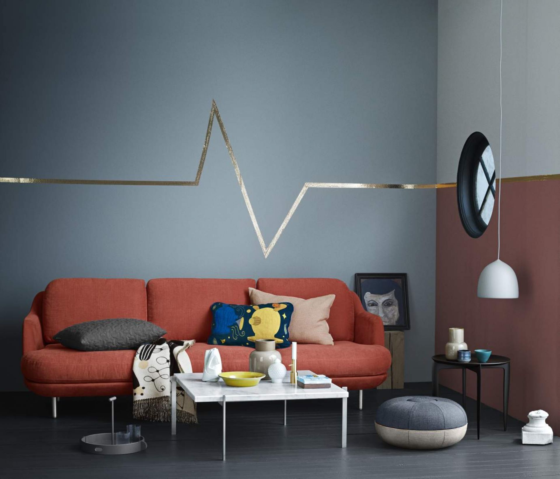 Wohnzimmer-Szene mit rotem Fritz Hansen Lune Sofa, Couchtisch, Kissen und Deko-Elementen vor einer blau-grauen Wand.