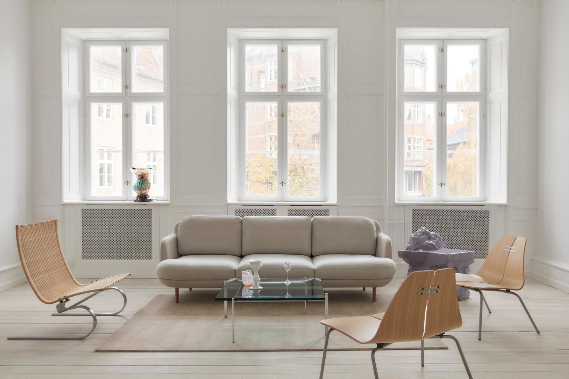 Heller Wohnraum mit PK23 Loungesessel von Fritz Hansen, Sofa, Glastisch und Design-Stühlen vor großen Fenstern.