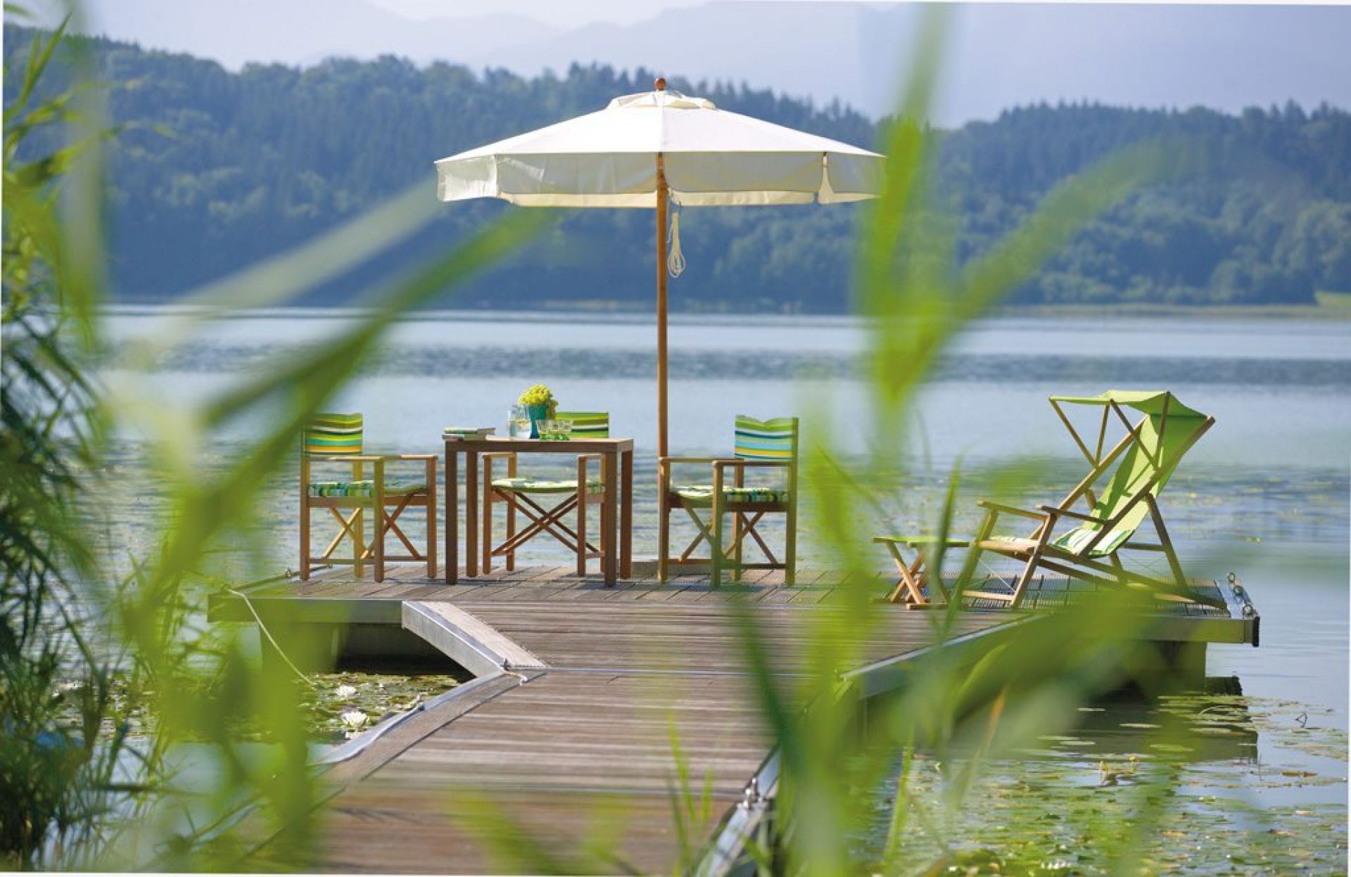 Weishäupl Cabin Outdoor Liegestuhl Deluxe am See, mit Tisch, Stühlen und Sonnenschirm.