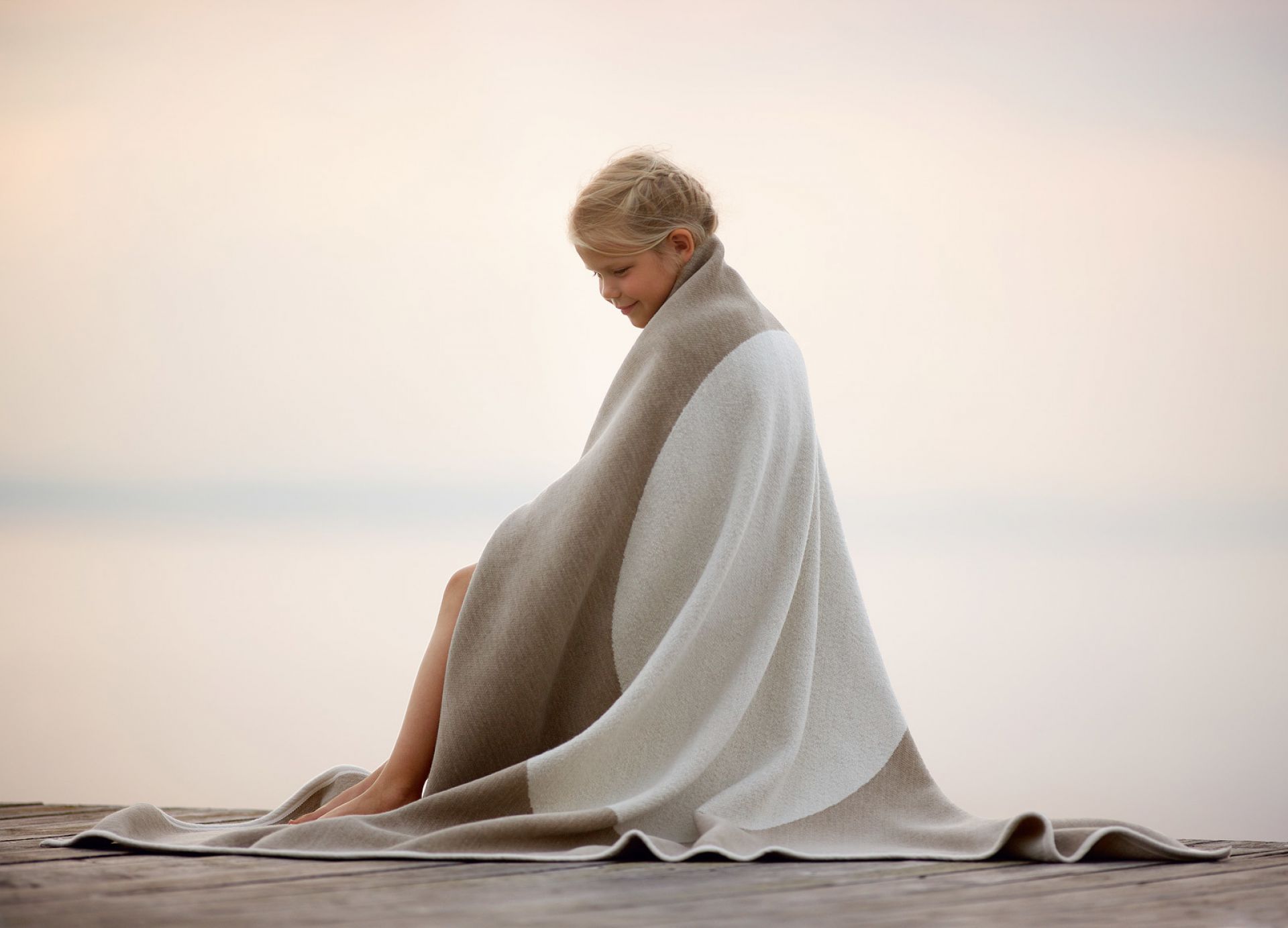 Mädchen gehüllt in Pappelina Vera Wolldecke beige-weiß, kuschelig weiche Wohndecke aus Wolle für gemütliche Stunden.