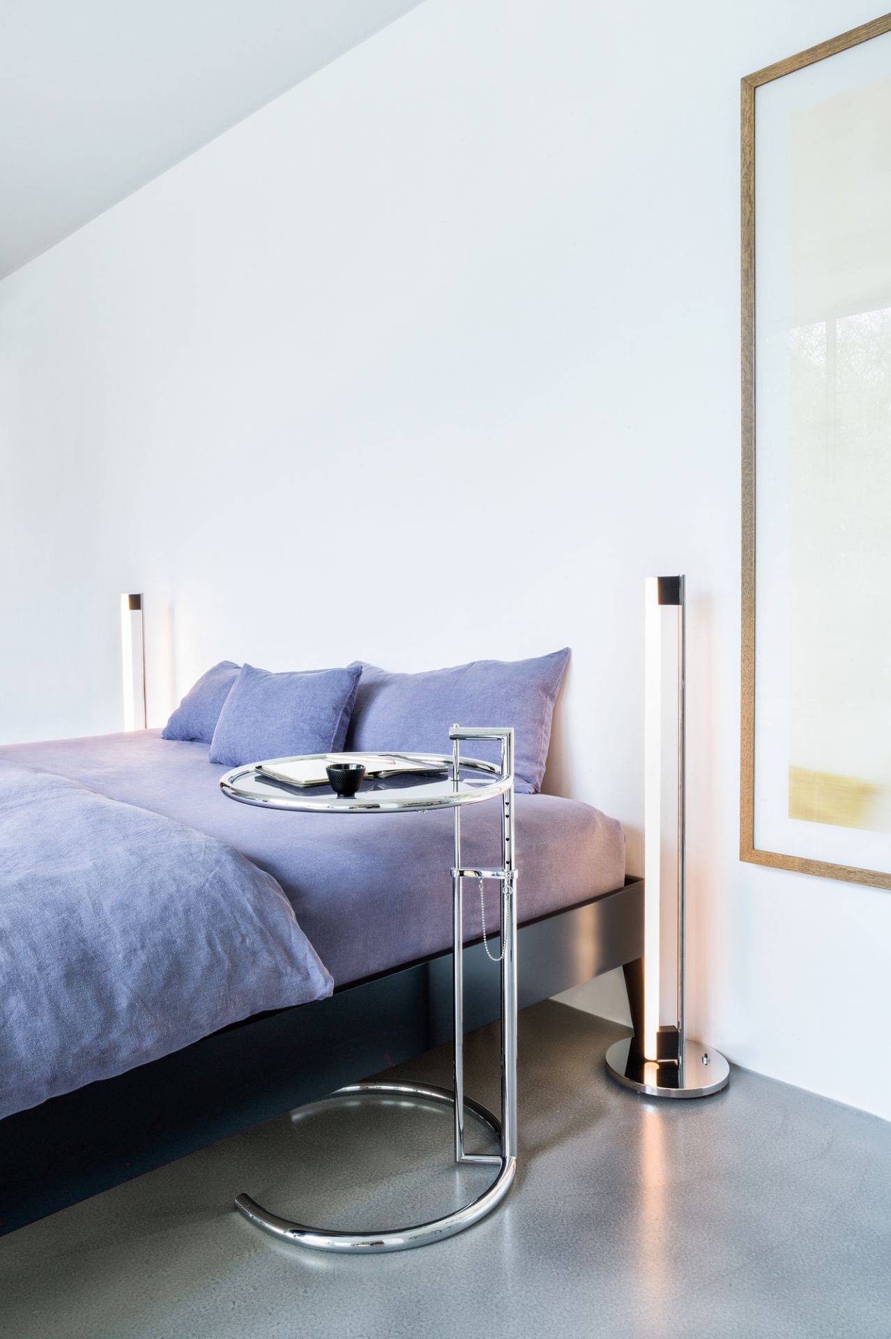 ClassiCon E1027 Beistelltisch, höhenverstellbar, Chromgestell neben einem Bett mit Notizbuch und Tasse.