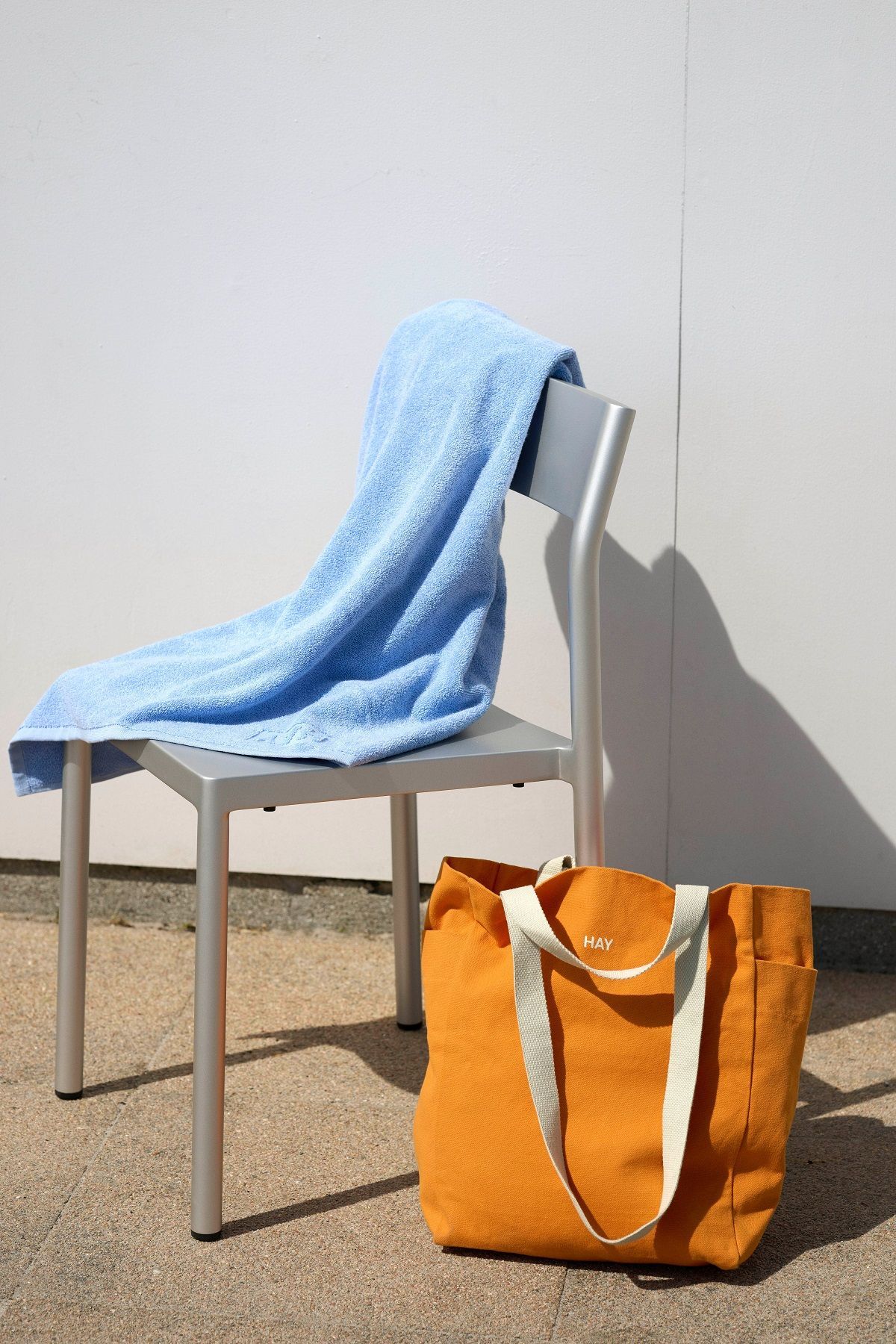 Orangefarbene Hay Everyday Tote Bag aus Stoff mit weißen Trägern neben einem Stuhl mit blauem Handtuch.