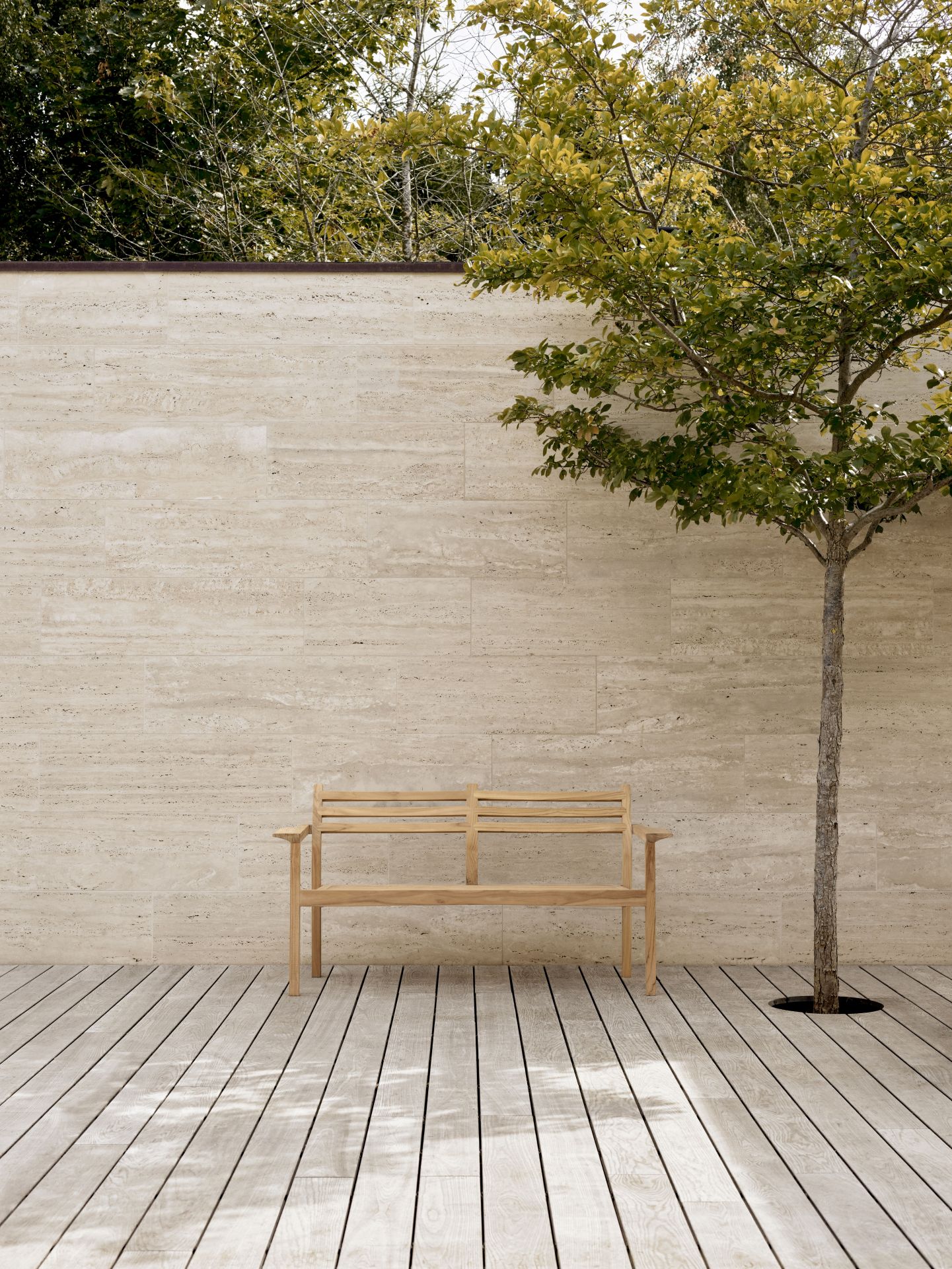 AH701 Carl Hansen Teak Gartenbank auf Holzterrasse vor Travertinwand und Baum.