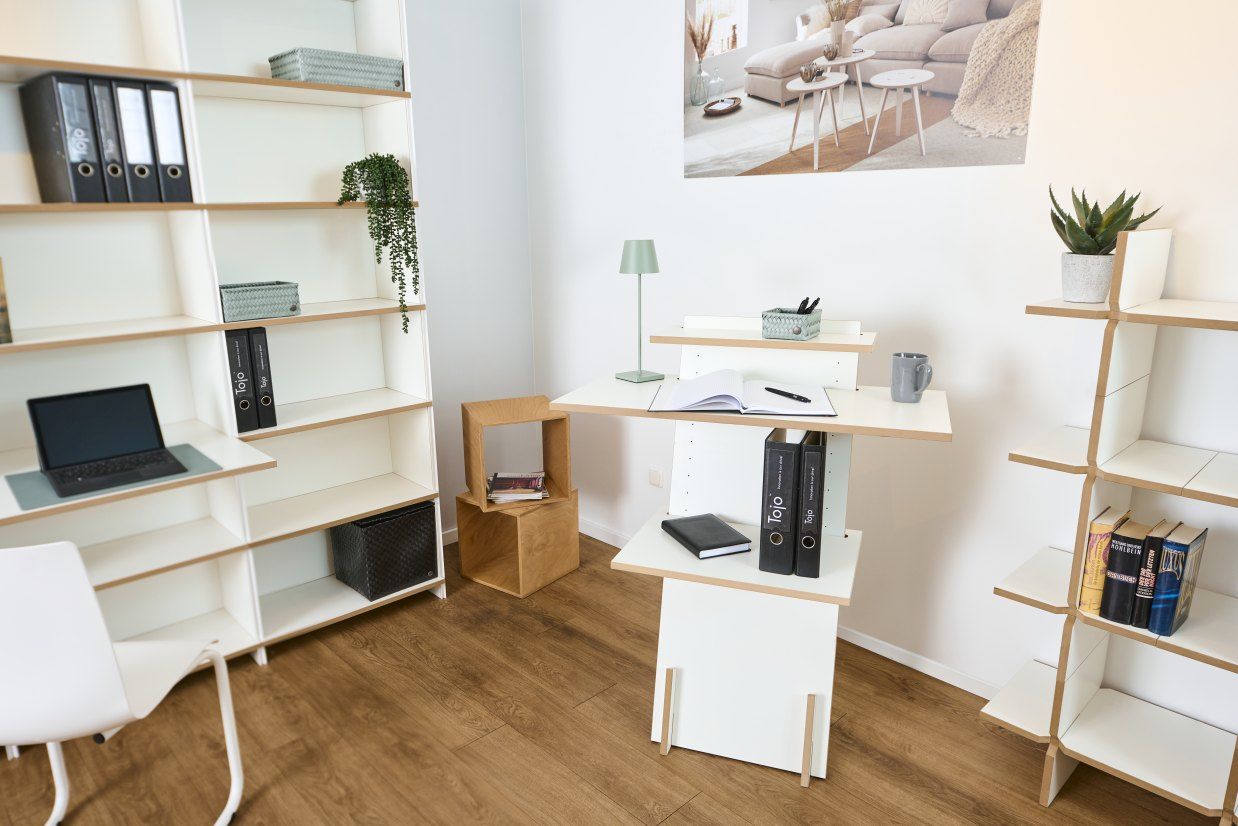 Helles Büro mit Tojo Stehpult aus Holz und weißen Regalen, Laptop, Ordnern und Dekoration.