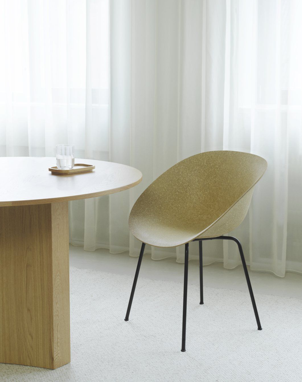 Normann Copenhagen Mat Armchair mit Stahlbeinen neben Holztisch und Glas Wasser vor transparenten Vorhängen.