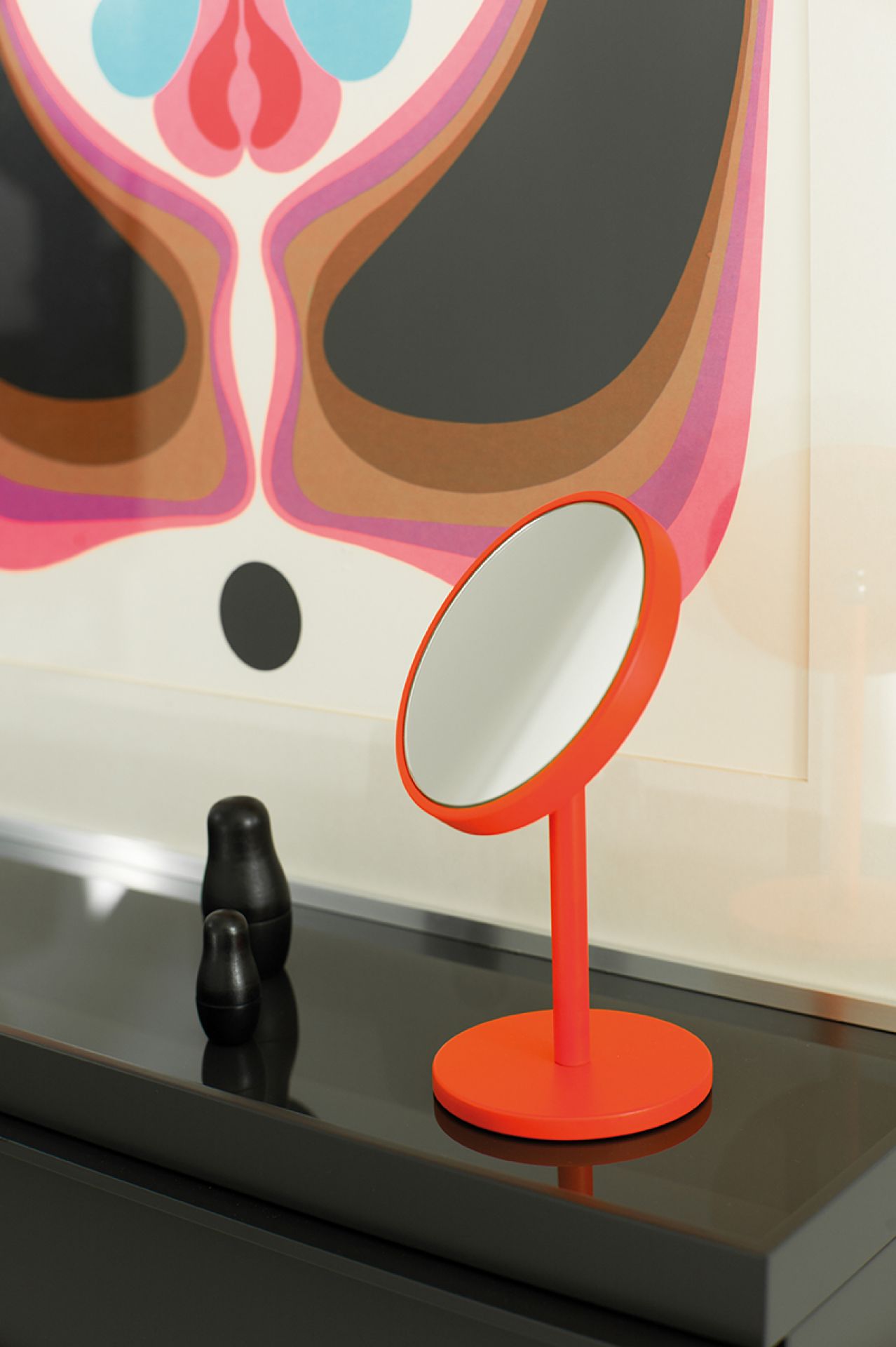 Oranger Schönbuch Beauty Schminkspiegel auf schwarzer Kommode, daneben Matroschka-Puppen und ein abstraktes Wandbild.