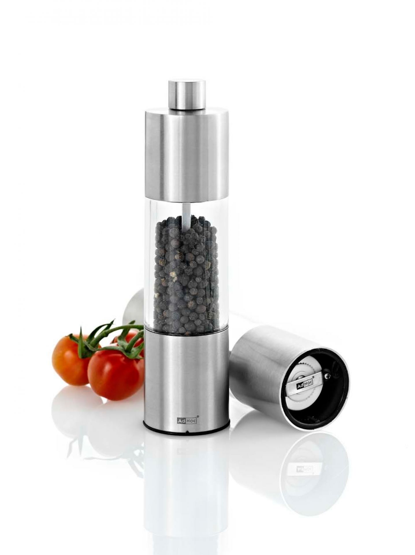 AdHoc Pfeffermühle Classic Medium aus Edelstahl und Acrylglas, gefüllt mit Pfefferkörnern, neben Tomaten.