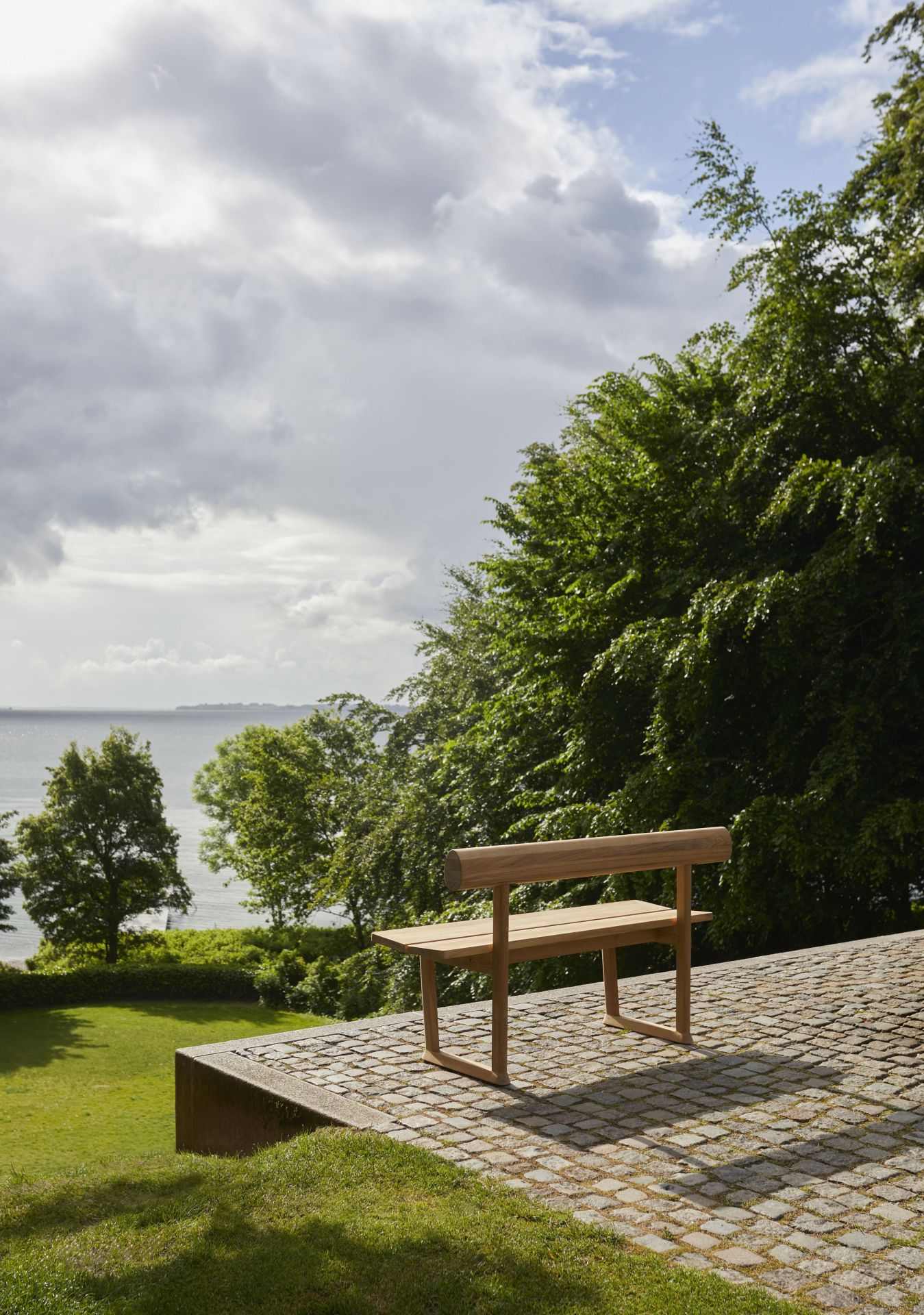 Banco Bank Teak Outdoor Skagerak by Fritz Hansen auf gepflasterter Fläche mit Blick auf Meer und Bäume.
